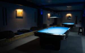 Dimly lit pool hall with two blue billiards tables under hanging lights