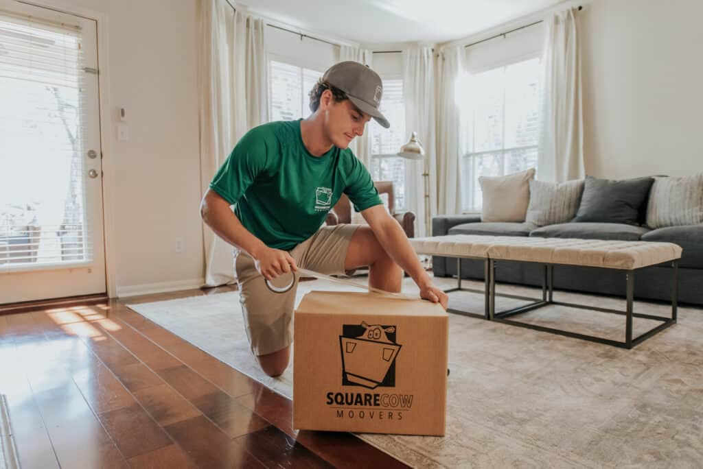 Mover kneeling on the floor taping a moving box in a bright living room
