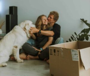 A man and woman sit on the floor with a dog, next to a box