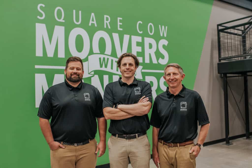 Three men in Square Cow Movers shirts pose in front of a green company wall