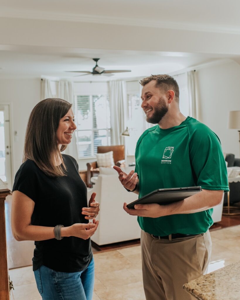 A "Square Cow Movers" employee speaks with a customer in the living room, both smiling.