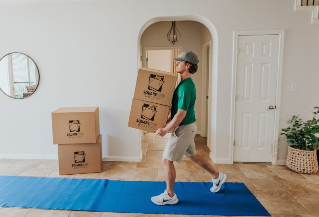 A mover carries two stacked boxes into a home while other boxes are nearby.