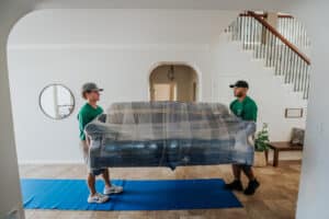 Two movers carrying a couch covered in plastic wrap into a home