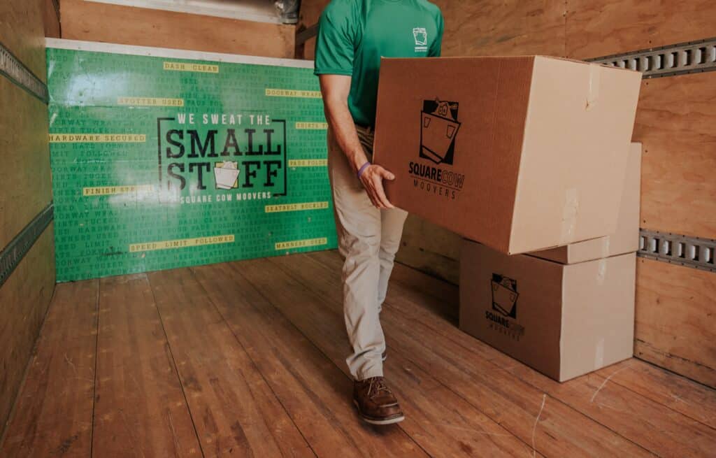 A moving crew member carries a box in a truck with a "Square Cow Movers" logo and a green wall that reads "We Sweat the Small Stuff."
