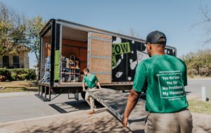 Two movers unloading equipment from a truck, one with a dolly and the other with a ramp.