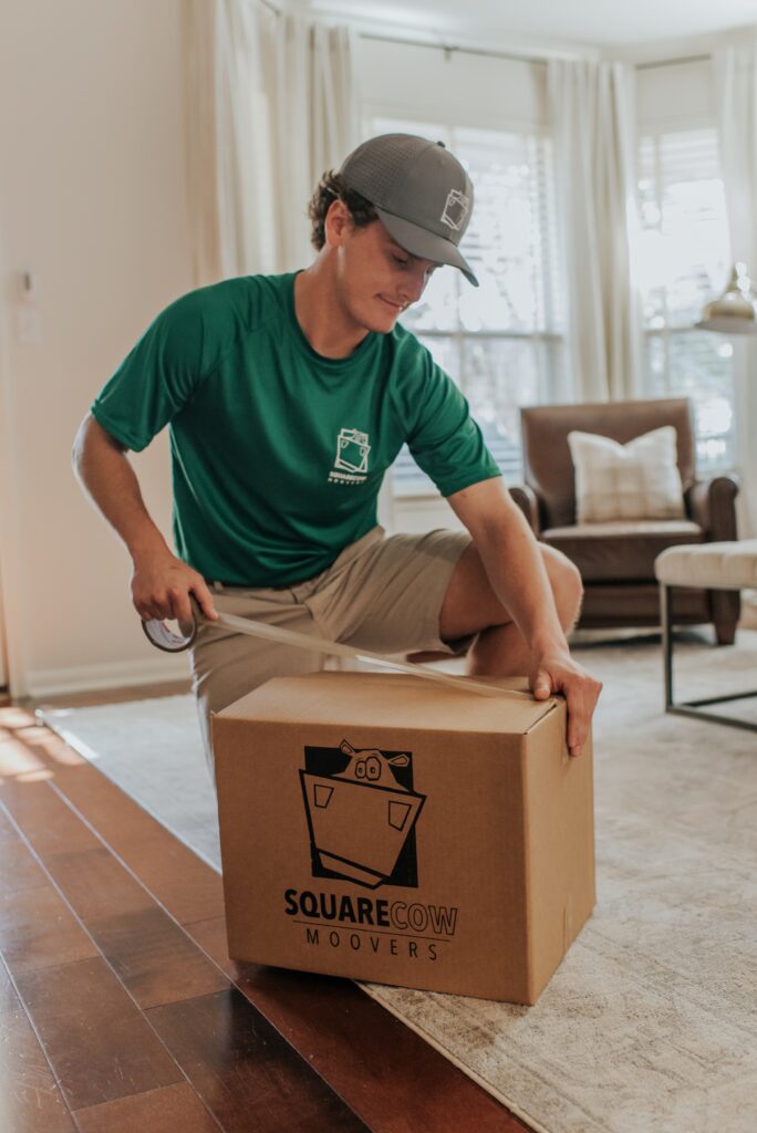 A mover in a green shirt and cap tapes up a cardboard box labeled "Square Cow Moovers" inside a living room.