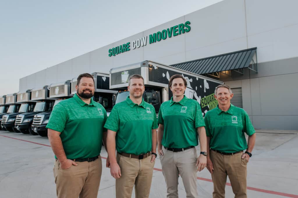 Four men in green shirts stand in front of a Square Cow Movers building with moving trucks.